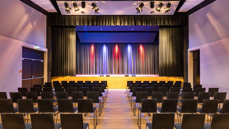 Sconarium - Event hall, © Wiener Alpen, Christian Kremsl An empty event hall with rows of chairs and a stage with curtains and colorful lighting.