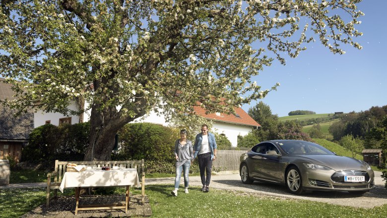 E-mobility, © Wiener Alpen A couple walks next to a parked electric car under a blossoming tree in a rural garden.