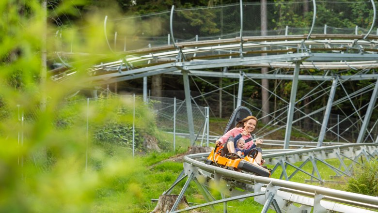 Sommerrodelbahn, © Erlebnisarena St. Corona Eine Frau und ein Kind fahren lachend auf einer Sommerrodelbahn durch eine grüne Landschaft.