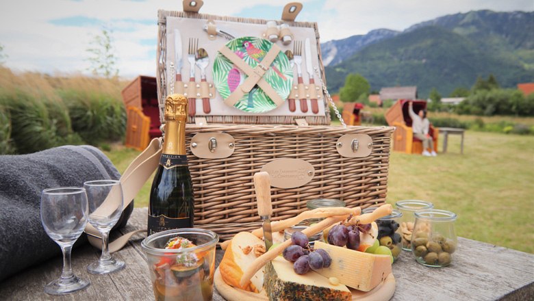 Picknick Korb, © Schloss Wartholz - Nusterer Picknickkorb mit Käse, Trauben und Sektflasche auf einem Tisch in einer ländlichen Umgebung mit Bergen im Hintergrund.