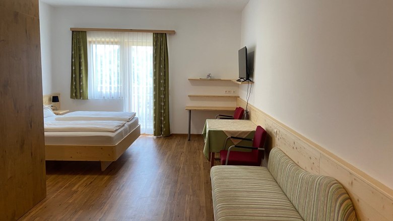 Familienzimmer, © Wiener Alpen Ein modernes Hotelzimmer mit Doppelbett, Sofa, Tisch und Stühlen. Ein Fernseher hängt an der Wand. Vorhänge an einem Fenster lassen Tageslicht herein.