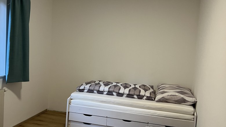 Zweibettzimmer, © Andrea Tauchner Ein einfach eingerichtetes Zweibettzimmer mit Holzboden, zwei Einzelbetten und einem Fenster mit blauen Vorhängen.