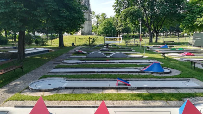 Minigolf course, © Minigolf Wiener Neustadt Minigolf course with several courses and obstacles, surrounded by trees and a high tower in the background.
