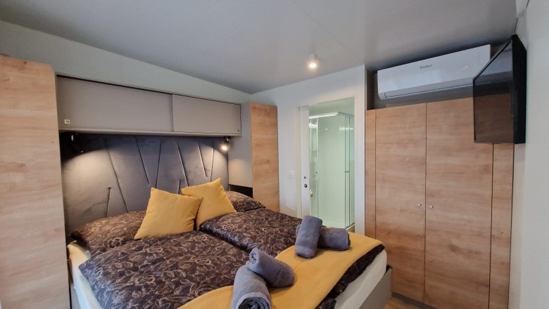 Doppelbett Johannesbachklamm Chalet, © Johannesbachklamm Chalet Innenansicht eines Schlafzimmers mit Doppelbett, gelben Kissen und Handtüchern, Schrank, Klimaanlage und Blick ins Badezimmer.