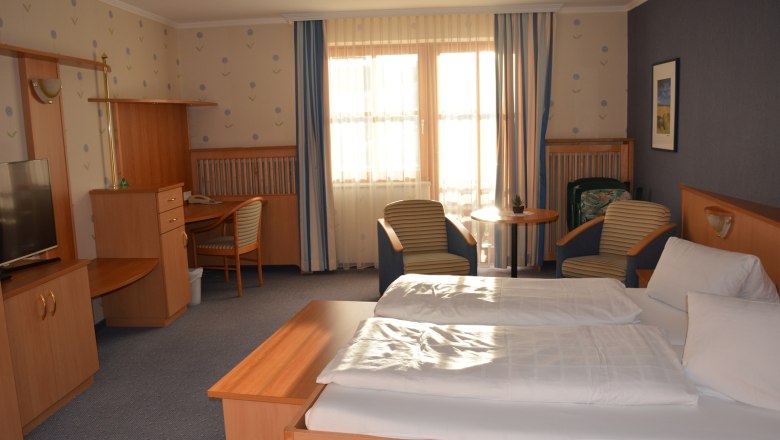 Doppelzimmer, © Michele Unger Ein gemütliches Hotelzimmer mit Doppelbett, Schreibtisch, Fernseher und zwei Sesseln vor einem Fenster mit Vorhängen.