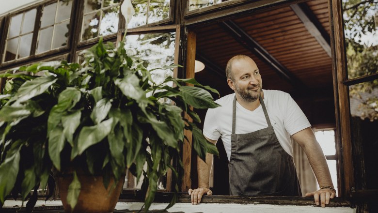 Landlord Andreas Unger looks after his guests, © Niederösterreich Werbung/Sophie Menegaldo A man in an apron leans out of a window, a plant in the foreground.