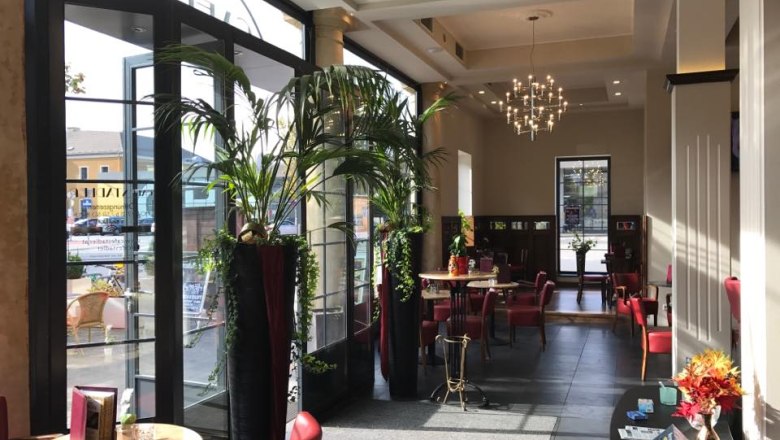 Café Stadler, © Café Stadler Innenansicht eines eleganten Cafés mit roten Stühlen, großen Fenstern und Pflanzen.