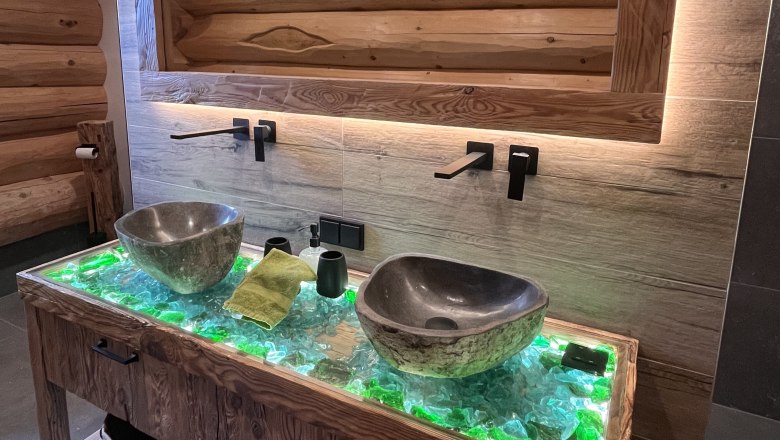 Badezimmer, © RaxChalet Modernes Badezimmer mit zwei steinernen Waschbecken auf einem beleuchteten Holztisch.