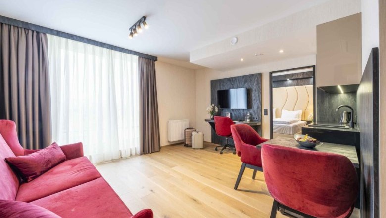 PLAZA Residence Longstay, © PLAZA Residence Wiener Neustadt Modernes Hotelzimmer mit rotem Sofa, Schreibtisch und Fernseher.