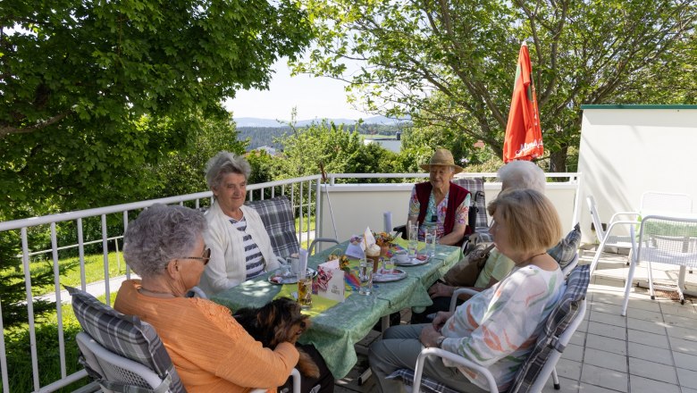 Shared conversations on the terrace, © ÖJAB Waldpension Elderly people sit around a table on a terrace, surrounded by trees and with a view of the countryside.