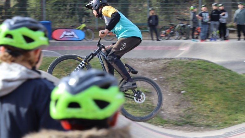 Pumptrack Neunkirchen, © Stadtgemeinde Neunkirchen Ein Radfahrer springt auf einem Pumptrack, während Zuschauer mit grünen Helmen zuschauen.