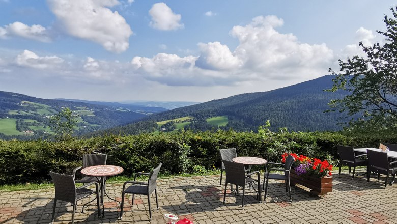 Traumhafter Ausblick auf der Terrasse, © Alpengasthof Fernblick, Fam. Pölzelbauer Terrasse mit Tischen und Stühlen, Blick auf grüne Hügel und blauen Himmel mit Wolken.