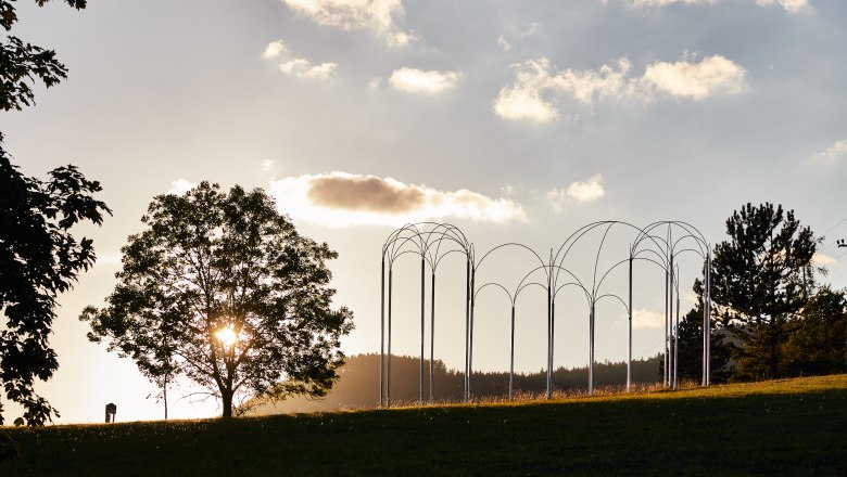 Gut Gasteil, © Nina Nagy Photography Ein Baum und eine Metallskulptur auf einer Wiese bei Sonnenuntergang.