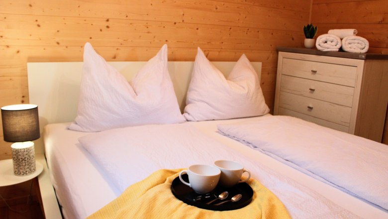 Schlafzimmer, © Fam. Auer Gemütliches Schlafzimmer mit Doppelbett, weißen Kissen und Decken, gelber Decke, Tablett mit Tassen, Nachttischlampe und Kommode mit Handtüchern.