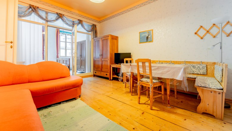 Wohnzimmer, © Wiener Alpen / Christian Kremsl Ein Wohnzimmer mit orangefarbenem Sofa, Holztisch und Stühlen, Holzboden und einem Schrank. Ein Bild hängt an der Wand.