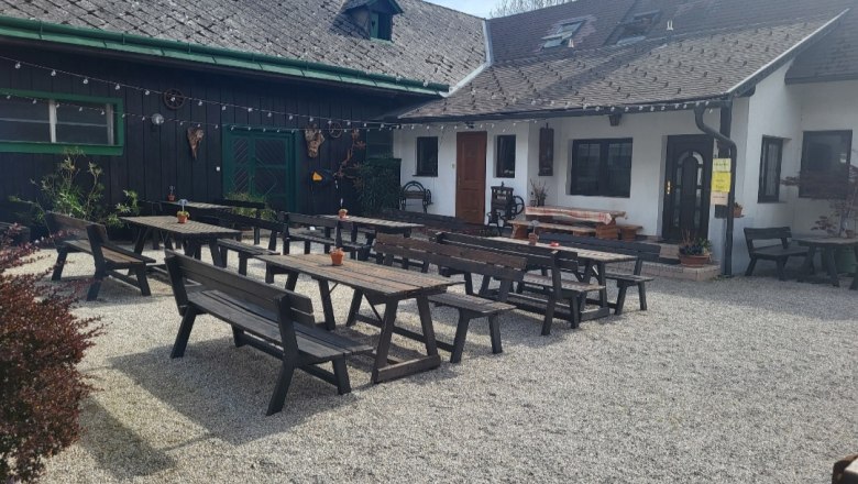 Leo-Hof Gastgarten, © Leo-Hof/Sandra Reinisch Ein Gastgarten mit Holzbänken und Tischen auf Kiesboden vor einem traditionellen Gebäude.