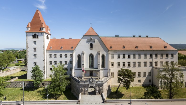 Militärakademie Westtor, © Wiener Alpen, Foto: Franz Zwickl Historisches Gebäude mit rotem Dach und Turm, umgeben von Bäumen und einer Straße im Vordergrund.