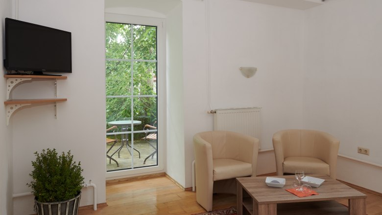 Wohnbereich, © Landhaus Puchbergerhof by smc Wohnzimmer mit zwei beigen Sesseln, Holztisch, Fernseher und Balkon mit Tisch und Stühlen.