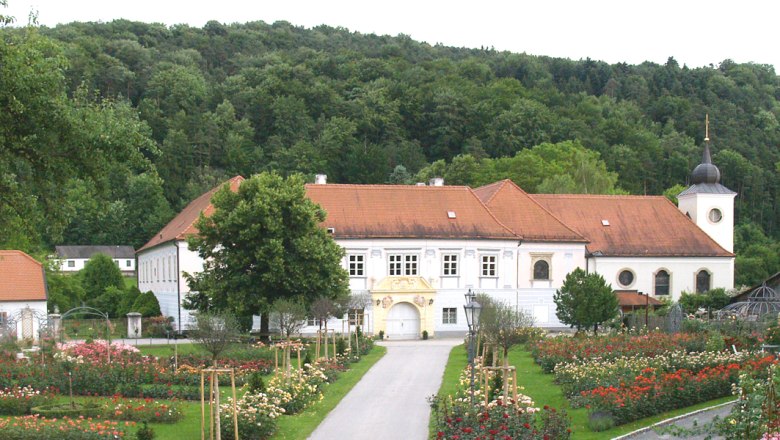 Rosengarten Pitten, © Rosengartenverein Pitten Ein historisches Gebäude mit rotem Dach, umgeben von einem Rosengarten und Bäumen, im Hintergrund ein bewaldeter Hügel.
