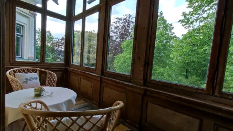 Veranda, © Villa Petterhof Veranda mit Tisch und zwei Stühlen, umgeben von großen Fenstern mit Blick auf grüne Bäume.