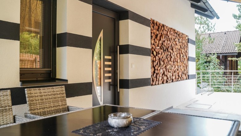 Eingang Ferienhaus Waxeneck, © Michaela Narath Eingang eines Ferienhauses mit moderner Tür, gestapeltem Holz an der Wand und einem Tisch mit Stühlen im Vordergrund.