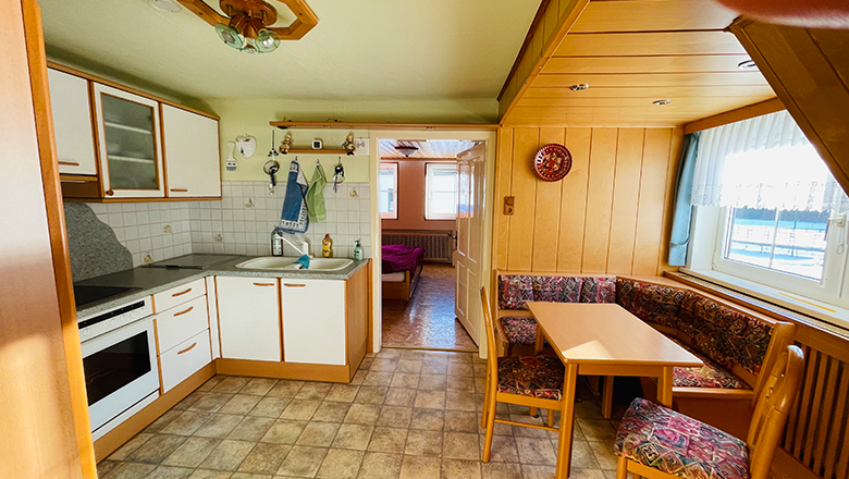 Kitchen, © Angelika Burger Cozy kitchen with corner bench, table and chairs, light wooden furniture, tiled floor, window with curtains, view into adjoining room.
