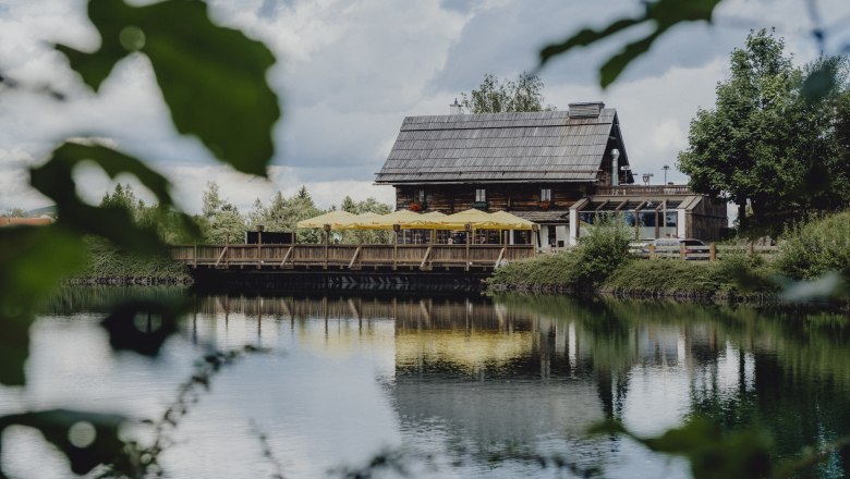 Am Fuße des Hirschenkogels (Zauberberg Semmering), © Niederösterreich Werbung/Rita Newman Ein rustikales Holzhaus mit gelben Sonnenschirmen spiegelt sich in einem ruhigen See, umgeben von grüner Natur.