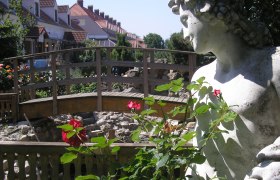 Zum Griechen, © Trifinopoulos Dimtiros Steinstatue neben einer Holzbrücke und roten Rosen im Vordergrund.