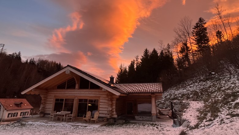 RaxChalet im Winter, © RaxChalet Ein Holzchalet im Winter mit schneebedecktem Boden und einem dramatischen, orangefarbenen Himmel im Hintergrund.