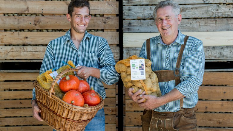 Potatoes, © Martina Siebenhandl Two smiling men in checked shirts hold a basket of pumpkins and a sack of potatoes in front of a wooden wall.