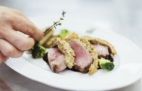 Rosa Hirschrücken mit Maronischupfnudeln, © Niederösterreich Werbung/David Schreiber Nahaufnahme eines rosa gebratenen Fleischgerichts mit Kräuterkruste, Brokkoli und einer Hand, die Thymian hinzufügt.