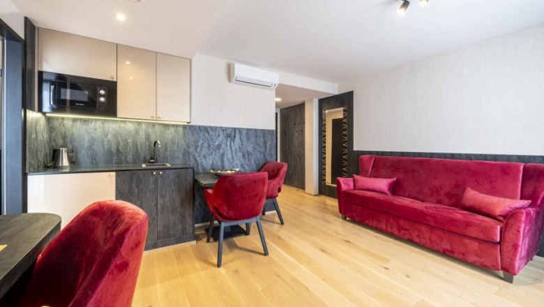 PLAZA Residence Longstay Zimmer, © PLAZA Residence Wiener Neustadt Modernes Apartment mit rotem Sofa und Stühlen, kleiner Küche und Holzboden.