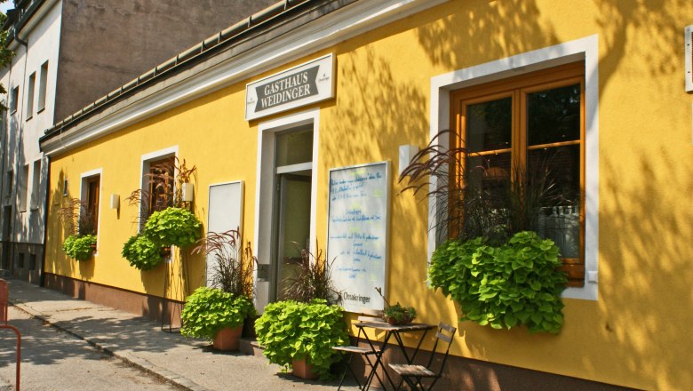 Weidinger Inn, © Wiener Alpen/Katrin Zeleny Yellow building with the inscription 'Gasthaus Weidinger', windows with flower boxes and a table with a chair in front of it.