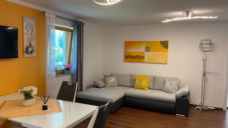 Wohnküche mit ausziehbarer Couch, © Wiener Alpen Wohnküche mit grauer ausziehbarer Couch, Esstisch und Dekorationen.