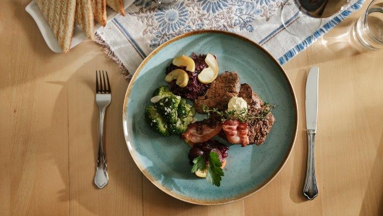Delicious food at the Raxalpenhof, © Sophie Menegaldo A plate of steak, broccoli, bacon and red cabbage on a table with cutlery and toast.