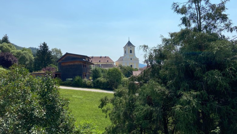 Aussicht vom Balkon Haus Köhler, © Wiener Alpen Blick auf eine Kirche und ein Holzhaus hinter Bäumen und Wiesen.