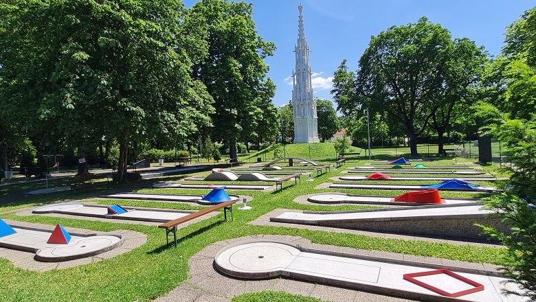 Minigolf Wiener Neustadt, © Minigolf Wiener Neustadt Minigolfplatz mit Hindernissen und einem hohen, weißen Turm im Hintergrund.