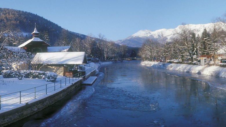 Payerbach entlang der Schwarza, © Wiener Alpen Winterlandschaft in Payerbach entlang der Schwarza mit schneebedeckten Bäumen und Bergen im Hintergrund.