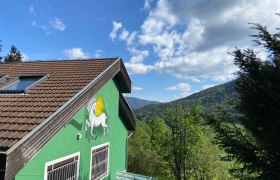 Einhornhaus, © Wieneralpen Grünes Haus mit Einhornbild an der Wand, umgeben von Bäumen und Bergen unter blauem Himmel.
