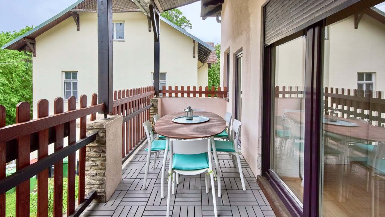 Top 2 - Balkon, © VP FeWo OG Ein Balkon mit Holztisch und Stühlen, umgeben von einem Holzzaun, mit Blick auf ein benachbartes Gebäude.