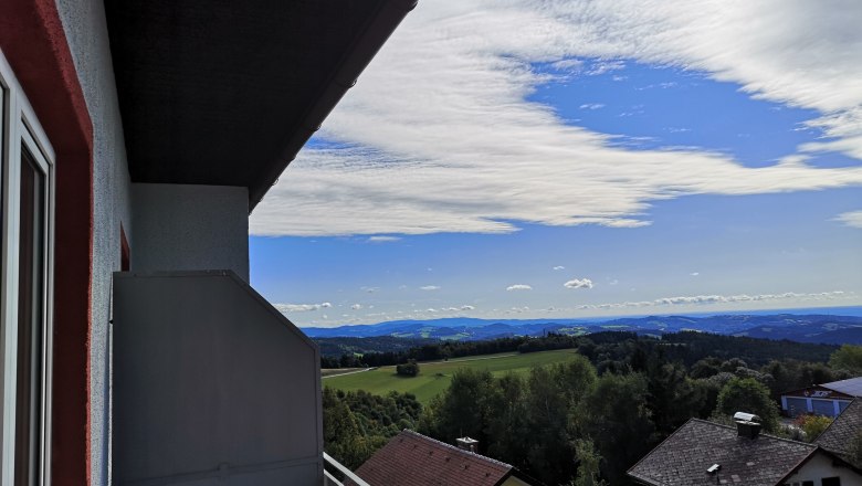 Balkon mit Ausblick, © Franz Thier Blick von einem Balkon auf eine Landschaft mit Häusern, Bäumen und Hügeln unter einem blauen Himmel mit Wolken.