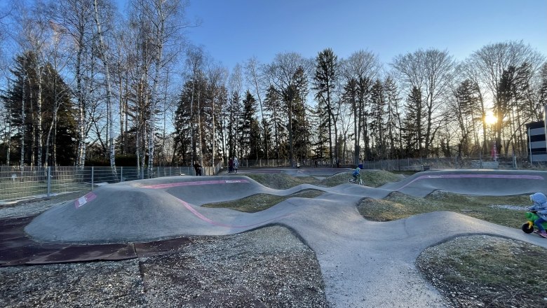 Pumptrack Neunkirchen, © Wiener Alpen/Lechner Pumptrack in Neunkirchen mit Kindern auf Fahrrädern und Laufrädern, umgeben von Bäumen bei Sonnenuntergang.