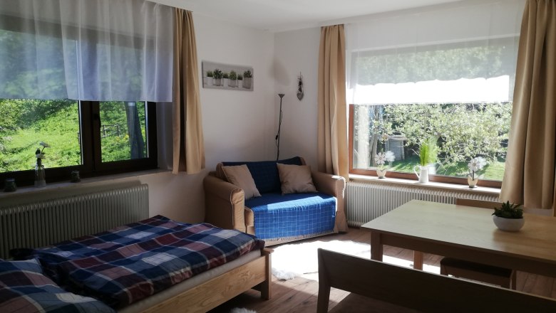 Ferienwohnung, © Familie Weibels Helles Wohnzimmer mit Bett, Sofa, Tisch und großen Fenstern mit Blick ins Grüne.