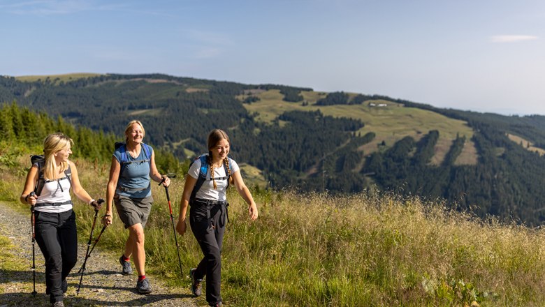 Tipp: 3 Hütten Schwaigen Tour, © Wiener Alpen, Christian Kremsl Drei Frauen wandern auf einem Bergpfad mit Wanderstöcken und Rucksäcken, umgeben von grünen Hügeln und Wäldern.