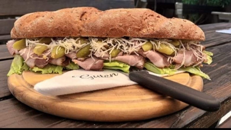 Leo-Hof Heurigenjause, © Leo-Hof/Sandra Reinisch Ein großes Sandwich mit Fleisch, Salat, Gurken und Käse auf einem Holzbrett, daneben ein Messer mit Serviette.