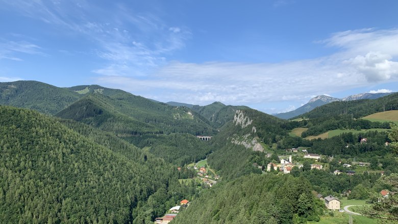 Breitenstein Blick, © Stefanie Gaulhofer Panoramablick auf eine grüne Berglandschaft mit einem kleinen Dorf und einem Viadukt.