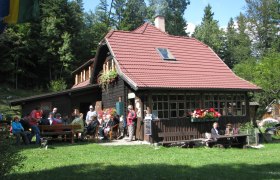 Turmsteighütte, © Naturpark Hohe Wand, Foto Privat Eine Berghütte mit rotem Dach, umgeben von Wald, mit Menschen, die draußen sitzen.