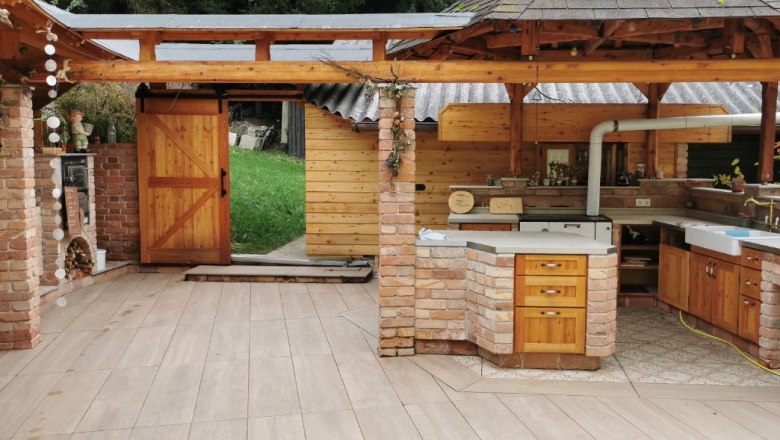 Mei Kuchl, © Anita Brem-Hofer Offene Außenküche aus Ziegelsteinen mit Holzmöbeln und Holzdach und mit Zugang zu einer grünen Wiese.