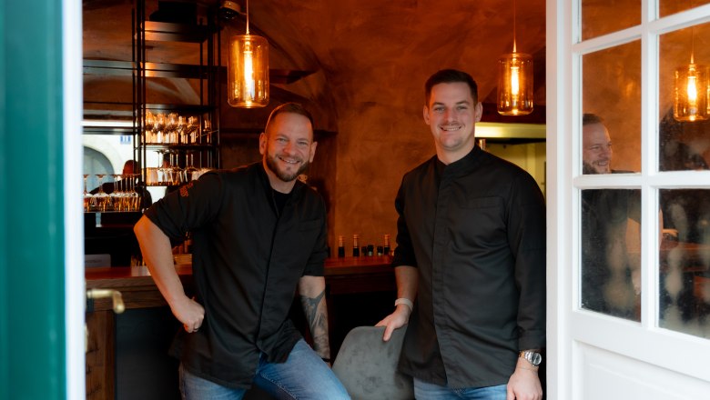 Die Gastgeber Daniel Alcántara García und William Merl, © Philipp Blickfang photography Zwei Männer in schwarzer Kleidung stehen lächelnd in einem stilvoll beleuchteten Raum mit Bar.