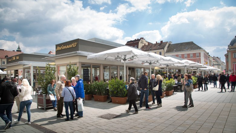 Marienmarkt, © WNSKS Menschen auf einem belebten Marktplatz mit Cafés und Geschäften unter Sonnenschirmen.
